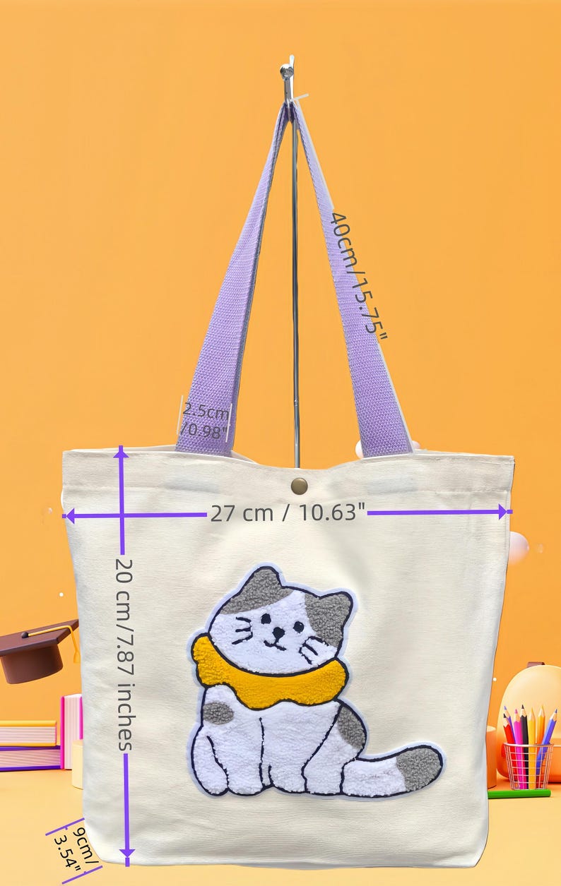 May include: Cream-colored tote bag with a cat design. The bag has purple handles and a button closure. The cat is white and gray with a yellow collar. The bag is 10.63 inches wide, 7.87 inches tall, and 3.54 inches deep.