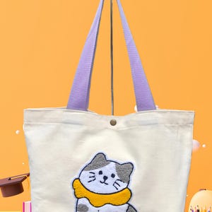 May include: Off-white canvas tote bag with a cartoon cat applique. The cat has gray and white fur with a yellow collar. The bag has purple handles and a button closure. The background is a gradient of orange and yellow.