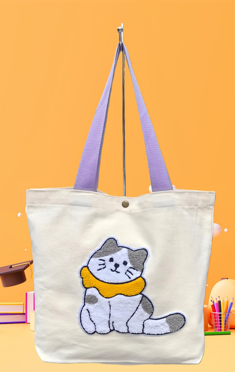 May include: Off-white canvas tote bag with a cartoon cat applique. The cat has gray and white fur with a yellow collar. The bag has purple handles and a button closure. The background is a gradient of orange and yellow.