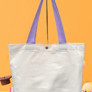 May include: A white tote bag with lavender-colored handles. The bag has a button closure and is made of a sturdy material. The tote bag is suitable for carrying books, groceries, or other items. The background is a solid orange color.