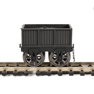 May include: A black model train coal car with two sets of wheels, sitting on a section of model train tracks. The coal car has a rectangular body with vertical slats and is filled with coal. The image includes a blue circle with the text "SAM'S TRAINS".