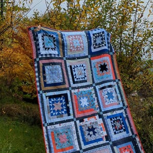 Pode incluir: Um cobertor de retalhos com um esquema de cores azul, branco e vermelho. O cobertor é feito de quadrados com padrões diferentes, incluindo estrelas, listras e padrões xadrez. O cobertor está pendurado em um varal em um jardim.