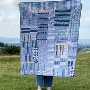 May include: A patchwork quilt made from blue and white fabric with a variety of patterns. The quilt is held up in front of a green grassy field and a blue sky.