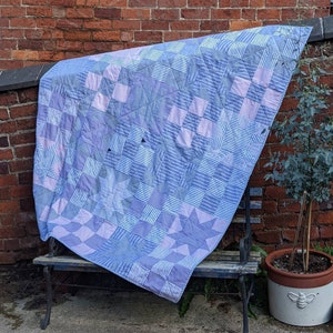Op de afbeelding: Een blauwe en roze patchworkdeken met een gestreept patroon is over een houten bank gelegd. De deken is gemaakt van verschillende stoffen en heeft een verscheidenheid aan kleuren en patronen.