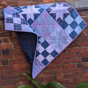 Op de afbeelding: Een patchworkdeken gemaakt van hergebruikt stof in tinten blauw, roze en wit. De deken hangt aan een bakstenen muur en heeft verschillende geometrische patronen, waaronder sterren en vierkanten.
