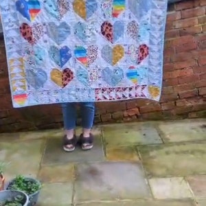 May include: A white quilt with a patchwork design featuring colourful hearts. The quilt is made of various fabrics with different patterns and textures. The quilt is being held up by a person, and the bottom of the quilt is visible.
