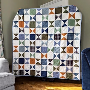 May include: A patchwork quilt featuring a repeating star pattern composed of squares and triangles. The design incorporates various fabric textures and patterns in shades of navy blue, olive green, burnt orange, and white, finished with a light blue border.