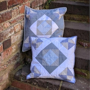 Peut inclure: Deux coussins carrés présentant un motif géométrique matelassé dans des tons de bleu, blanc et beige. Le design comprend des formes de losanges centraux entourés de petits triangles, avec des accents en tissu rayé bleu et blanc et des bords passepoilés.