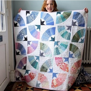 Op de afbeelding: Een quilt met een herhalend waaierpatroon in diverse tinten blauw, groen, roze en wit. Het ontwerp bestaat uit gebogen waaiervormige segmenten gerangschikt in blokken met stervormige centra, afgewerkt met gedetailleerd machinaal stikwerk.