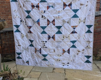 Puede incluir: Una colcha de patchwork cuadrada con un patrón repetitivo de pajaritas. El diseño incorpora bloques de tela blanca que se alternan con tela lisa de color verde azulado, tela a cuadros y una tela blanca estampada con pequeñas siluetas de osos.