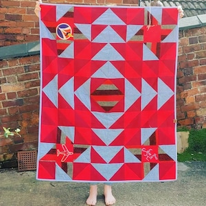 May include: A red and gray quilt with a geometric pattern. The quilt has a plane embroidered on it and the words "Son & Marge" in the bottom right corner.