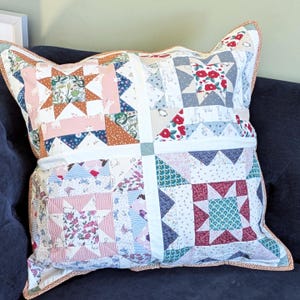 May include: A square throw pillow featuring a patchwork design with four distinct blocks separated by white sashing. The fabric patterns include floral prints, star motifs, and animal illustrations in shades of red, blue, pink, and green with orange trim.