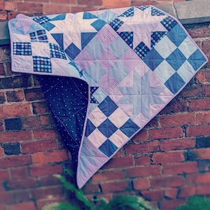 以下が含まれることがあります： 青、ピンク、白の配色でパッチワークされたキルト。キルトは正方形と星形でできており、レンガの壁にかけられています。