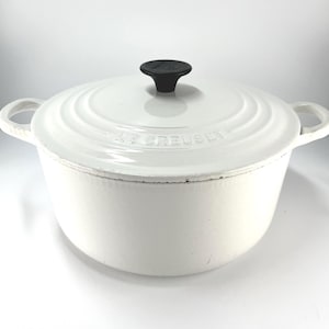 May include: A white enameled cast iron Dutch oven with a matching lid and a black phenolic knob. The lid features the embossed text LE CREUSET. The pot has two side handles and a smooth, glossy finish on the exterior surface of the round vessel.