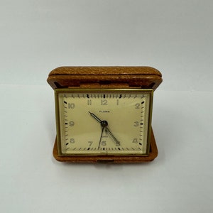 May include: A brown leather-covered travel alarm clock with a white face and black hands. The clock face reads "Florn" and has black Roman numeral hour markers.