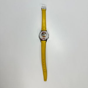 May include: A yellow leather wristwatch with a silver face featuring a cartoon character. The watch has a silver buckle.