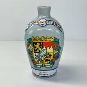 May include: A small, light blue ceramic bottle with a white label that reads "Deutsches Erzeugnis 40Vol.%". The bottle features a colorful coat of arms with a lion and a crown. The label also reads "Gruβ aus München".