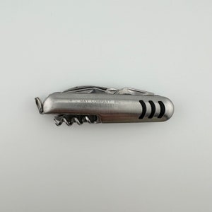 May include: A silver multi-tool pocket knife with a corkscrew, a W. L. May Company Inc. logo, and the text "Garden City, NY 11530".