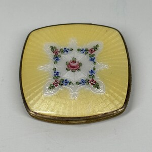 May include: A yellow enamel compact with a floral design. The compact has a white background with a pink rose in the center and blue and pink flowers around the edge. The compact is surrounded by a gold metal frame.