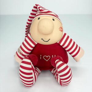 May include: A plush doll with a large nose, a red body, and striped red and white arms, legs, and hat. The doll has a smiling face, a kiss mark, and the words "i ♡ u" on its chest. The doll is sitting.