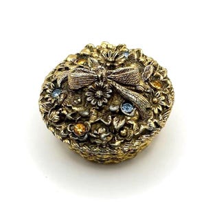 May include: A vintage, gold-toned jewelry box shaped like a floral basket. The box features a detailed design of flowers, leaves, and a bow, with accents of blue and orange gemstones. The box is round and has a textured surface.