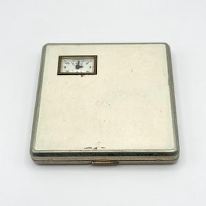 May include: A rectangular, silver-toned cigarette case with a small clock embedded in the upper left corner. The clock has a rectangular gold frame and a white face with black numerals. The case has a hinged closure and a slightly worn appearance.