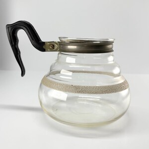 May include: Clear glass coffee pot with a black handle and a silver-colored metal rim. The pot has a decorative band of textured, light-colored material around the middle. The pot has a rounded shape with a series of horizontal ridges.