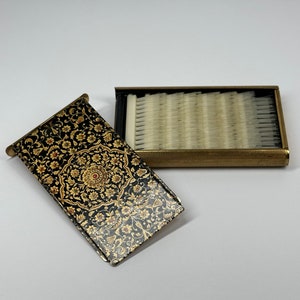 May include: A vintage black and gold compact with a floral design. The compact opens to reveal a row of white bristles.