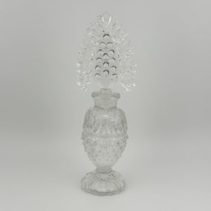 May include: A clear glass perfume bottle with a textured, ornate design. The bottle has a unique, decorative stopper shaped like a stylized tree.