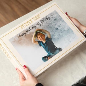 May include: A 1000-piece puzzle box with a watercolor-style image of a child holding a book over their head. The box is cream-colored with gold trim and the text "happy birth day my little boy".