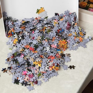 May include: A partially assembled jigsaw puzzle with many gray pieces scattered on a white surface. The puzzle pieces have various colors, including red, orange, yellow, and pink. The puzzle is in a white box.