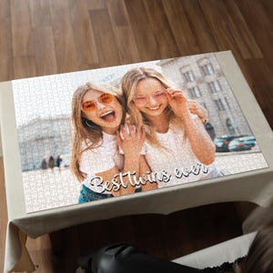 May include: A completed jigsaw puzzle featuring a photograph of two smiling women wearing sunglasses. The puzzle has the text "Best twins ever" in a cursive font. The puzzle is on a beige tablecloth.