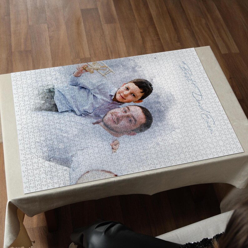 May include: A personalized jigsaw puzzle featuring a photograph of a man and a child. The puzzle is mostly white with a light-colored border and is displayed on a beige tablecloth. The image shows the faces of the man and child.