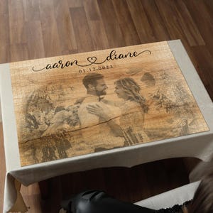 May include: A personalized wooden puzzle featuring a couple's photo. The puzzle has a rustic wood grain design with the names "aaron & diane" and the date "01.12.2023" printed on it. The puzzle is displayed on a white tablecloth.