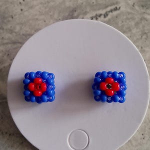Op de afbeelding: Een paar blauwe en rode kralen oorbellen. Elke oorbel is vierkant met een rode bloem in het midden.