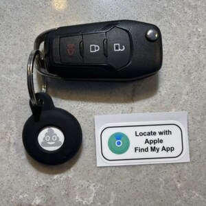 Puede incluir: Llave de coche negra con llavero, una etiqueta redonda negra con un emoji de caca y una pegatina blanca con el texto "Locate with Apple Find My App". La llave tiene tres botones.