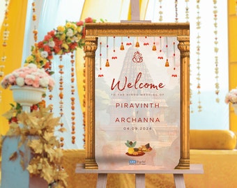 May include: A wooden easel displays a welcome sign for a Hindu wedding. The sign features a temple illustration, the names Piravinth and Archanna, and the date 04.09.2024. The sign is framed in gold and adorned with bells.