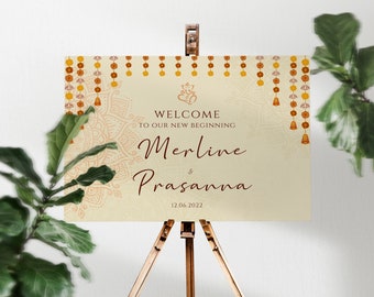 May include: Welcome sign with a cream background and gold and orange accents. The sign features a mandala design, a Hindu deity, and the text "Welcome to our new beginning, Merline & Prasanna, 12.06.2022".