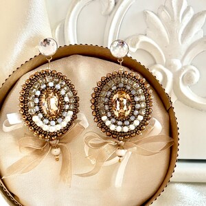 May include: A pair of round, beaded earrings featuring a central oval champagne-colored crystal, surrounded by concentric circles of bronze, white, and light blue beads. Each earring is accented with a sheer beige ribbon bow and a small bead drop.