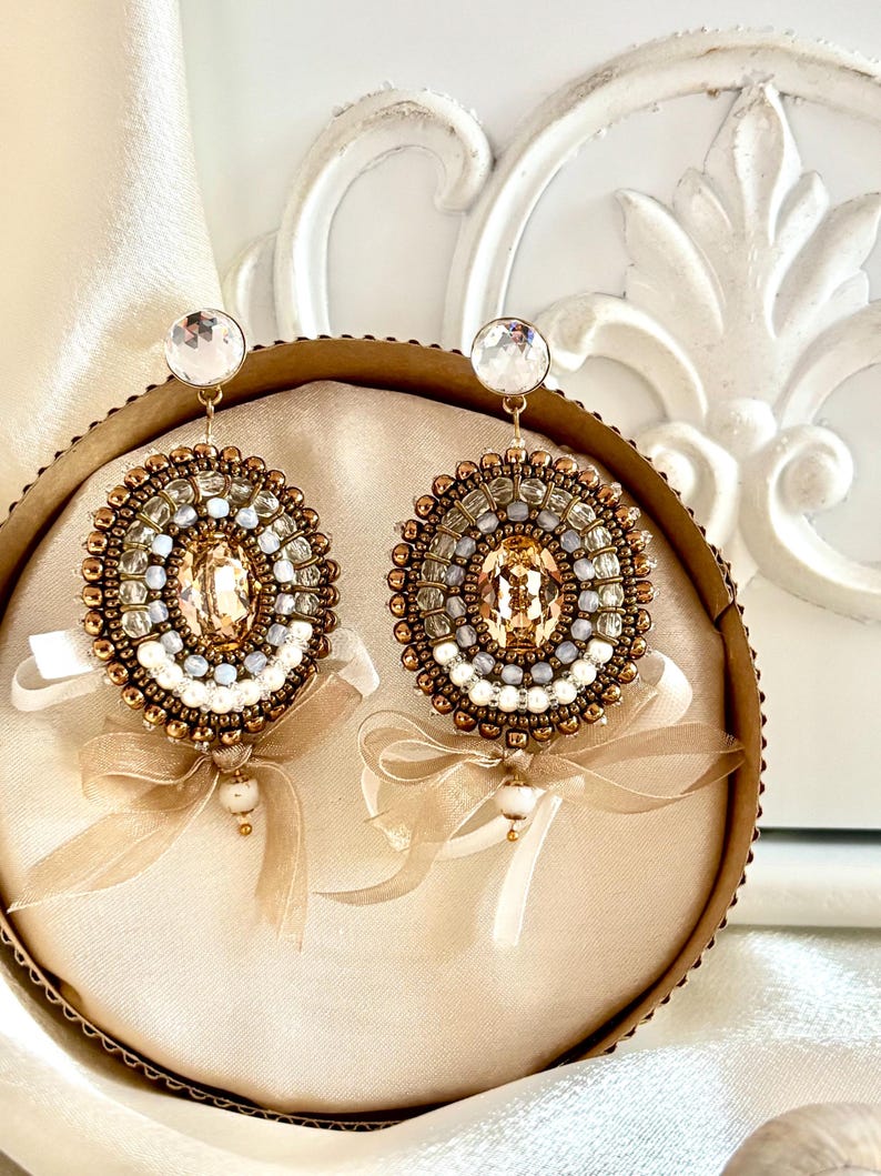 May include: A pair of round, beaded earrings featuring a central oval champagne-colored crystal, surrounded by concentric circles of bronze, white, and light blue beads. Each earring is accented with a sheer beige ribbon bow and a small bead drop.