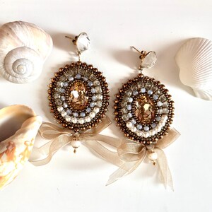 May include: A pair of circular beaded earrings featuring a central oval champagne-colored crystal surrounded by concentric rings of bronze, light blue, and white beads. Each earring is accented with a sheer beige ribbon bow and a small pearl.