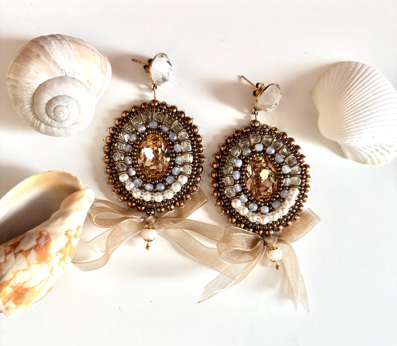 May include: A pair of circular beaded earrings featuring a central oval champagne-colored crystal surrounded by concentric rings of bronze, light blue, and white beads. Each earring is accented with a sheer beige ribbon bow and a small pearl.