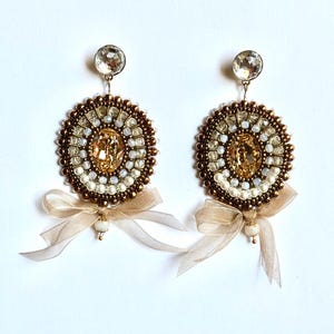 May include: A pair of circular beaded earrings featuring a central amber-colored crystal, surrounded by concentric rings of metallic bronze, white, and clear beads. Each earring is finished with a sheer beige ribbon bow and a small white bead.