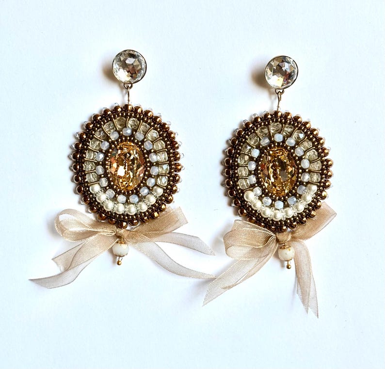 May include: A pair of circular beaded earrings featuring a central amber-colored crystal, surrounded by concentric rings of metallic bronze, white, and clear beads. Each earring is finished with a sheer beige ribbon bow and a small white bead.