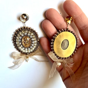 May include: A pair of circular earrings featuring a central amber-colored crystal surrounded by concentric rings of small metallic bronze and white beads. Each earring is topped with a clear faceted crystal and finished with a sheer beige ribbon bow.