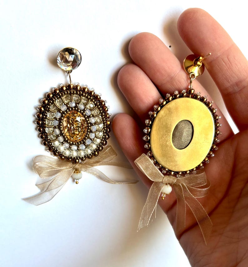 May include: A pair of circular earrings featuring a central amber-colored crystal surrounded by concentric rings of small metallic bronze and white beads. Each earring is topped with a clear faceted crystal and finished with a sheer beige ribbon bow.