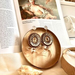 May include: A pair of round beaded earrings featuring a central champagne-colored crystal surrounded by concentric rings of small metallic bronze, silver, and white beads. Each earring is accented with a sheer, light beige ribbon bow at the base.
