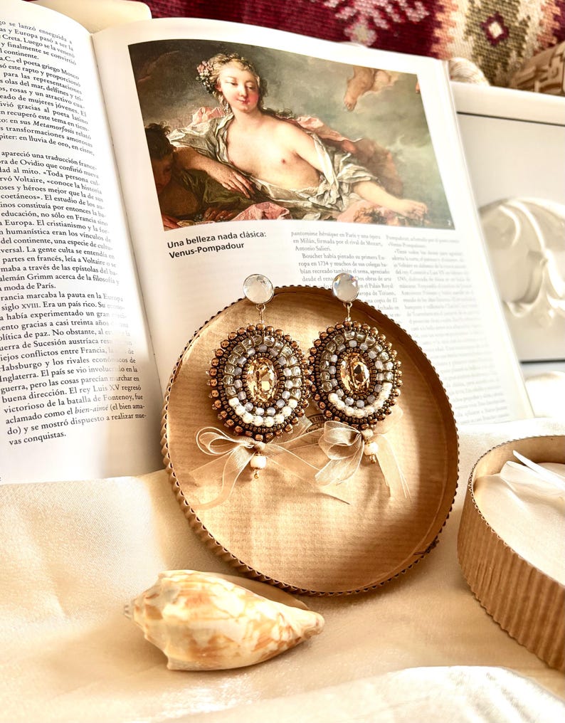 May include: A pair of round beaded earrings featuring a central champagne-colored crystal surrounded by concentric rings of small metallic bronze, silver, and white beads. Each earring is accented with a sheer, light beige ribbon bow at the base.