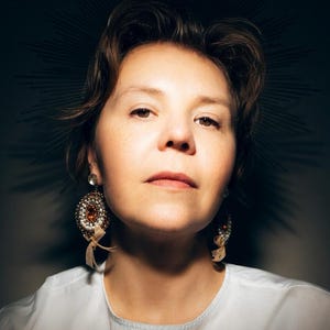 May include: A pair of ornate, circular drop earrings featuring a central amber-colored stone surrounded by intricate beadwork in shades of white, gold, and brown, finished with small, dangling beige ribbon accents attached to the bottom.
