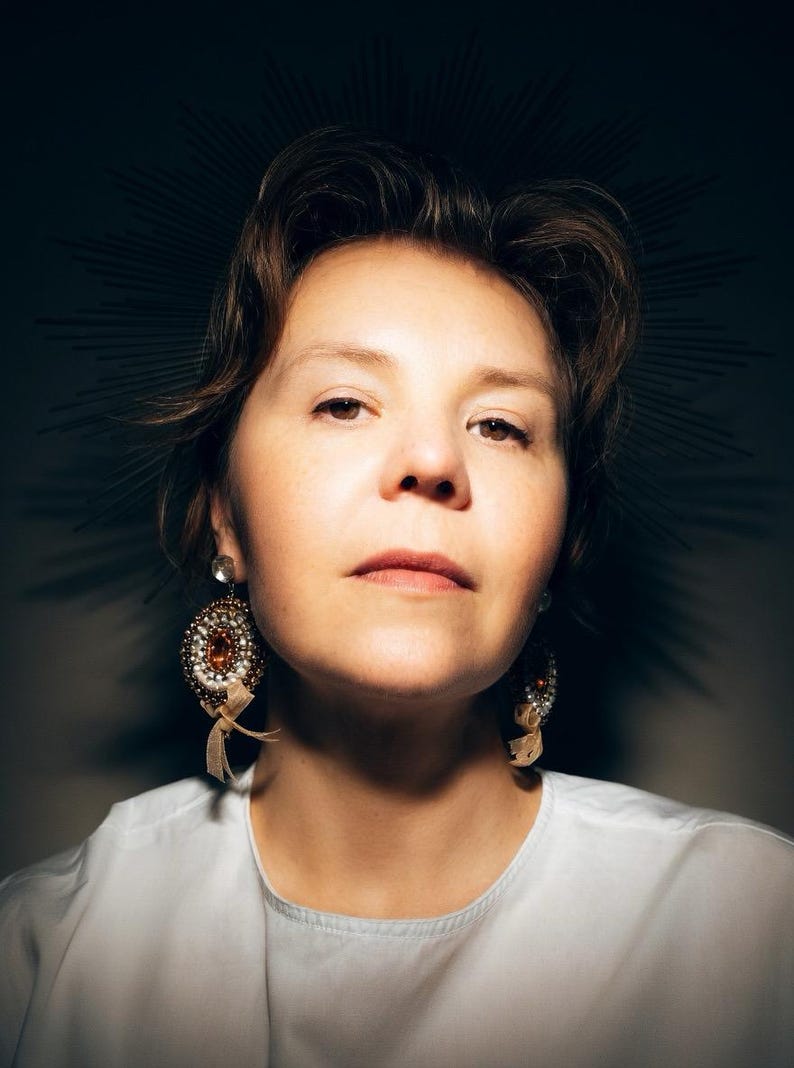 May include: A pair of ornate, circular drop earrings featuring a central amber-colored stone surrounded by intricate beadwork in shades of white, gold, and brown, finished with small, dangling beige ribbon accents attached to the bottom.